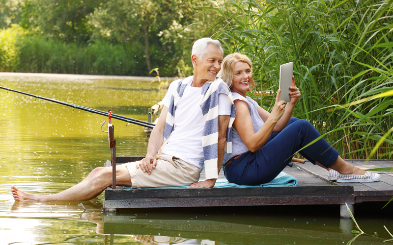 Portrait of elderly couple relaxing at nature. Senior woman and old man sitting back to back on the pier at lakeside while making self portrait with digital tablet.